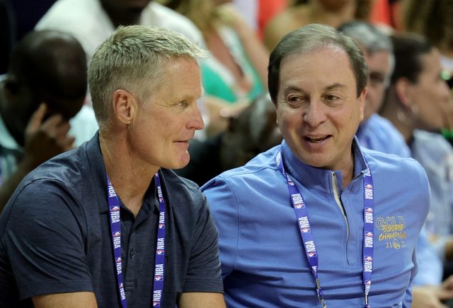 Two men wearing NBA lanyards are sitting close and engaged in conversation, with one wearing a dark shirt and the other a light blue ZIP-up.